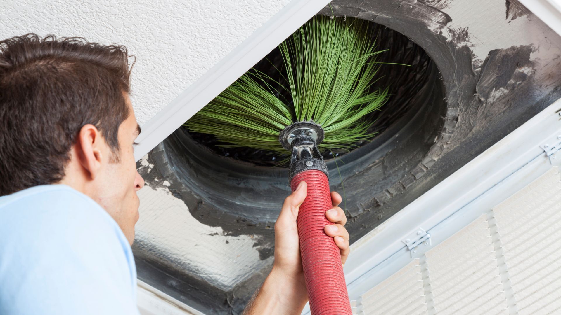 Close-up of a technician using a red-handled rotary chimney brush to clean a round flue opening, with green bristles spinning inside the duct to remove soot and debris.