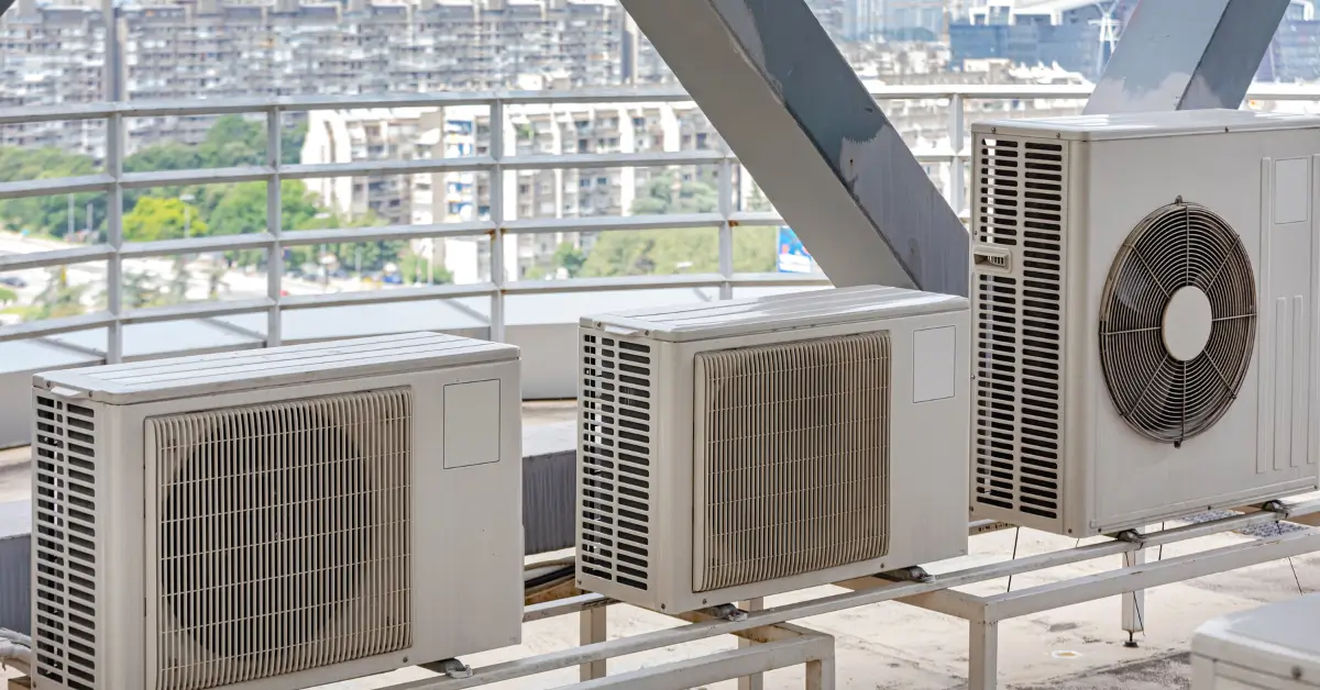 Three outdoor air conditioning condenser units installed on a rooftop platform, with a cityscape and high-rise buildings visible in the background.