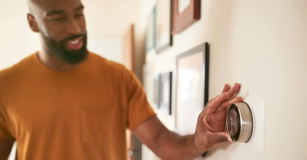 A man wearing a mustard-colored shirt adjusts a modern smart thermostat mounted on a wall, with framed pictures hanging in the background.