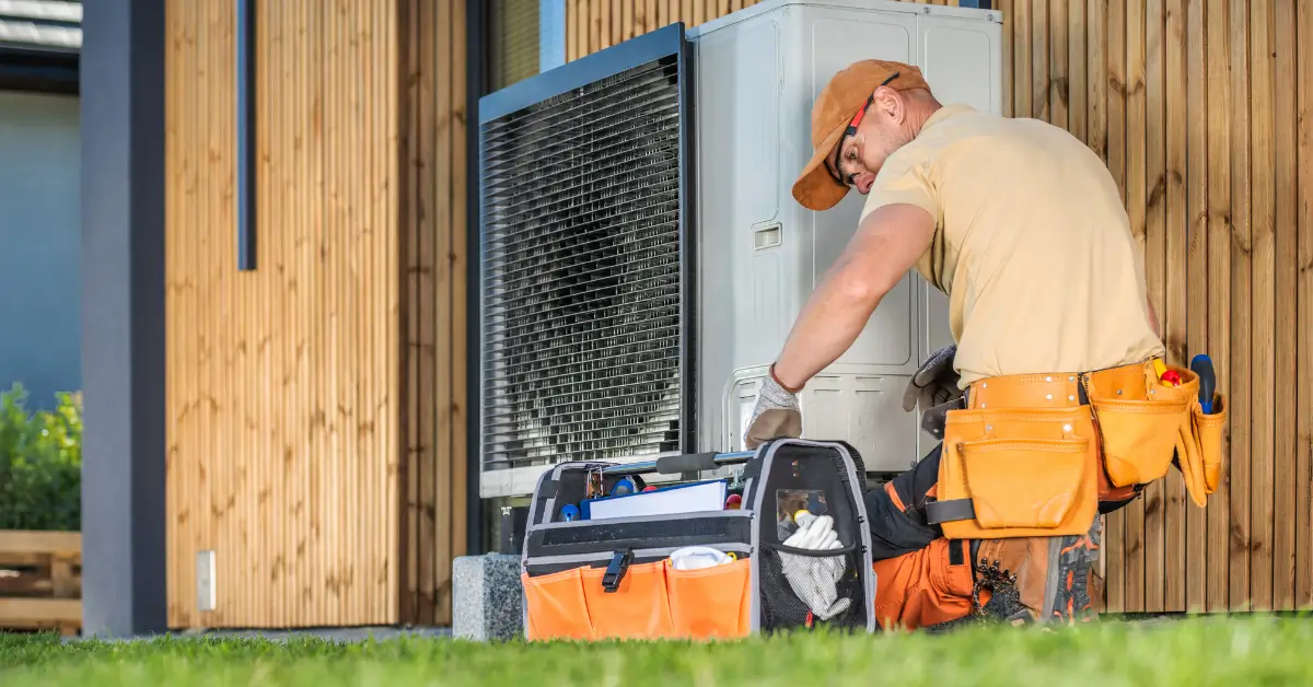 An HVAC technician wearing an orange cap, beige shirt, and tool belt works on an outdoor unit next to a modern wooden building, with a large open tool bag on the ground in front of him.