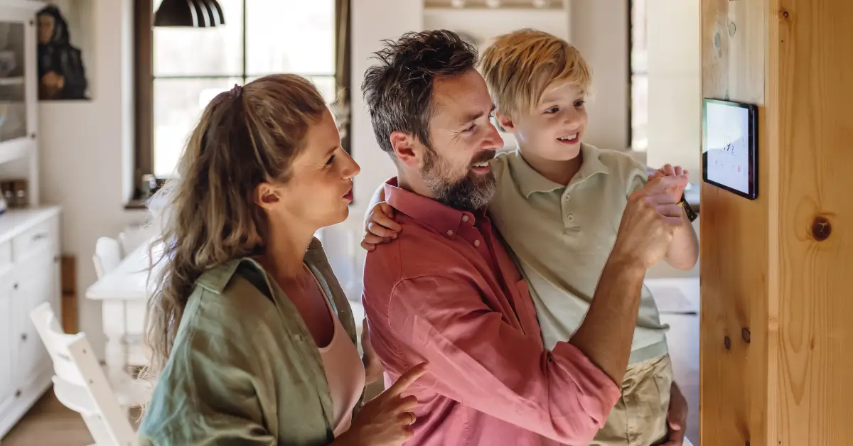 A smiling family of three stands in their home, with the father holding the young son as they adjust settings on a wall-mounted smart home control panel, while the mother looks on and points.