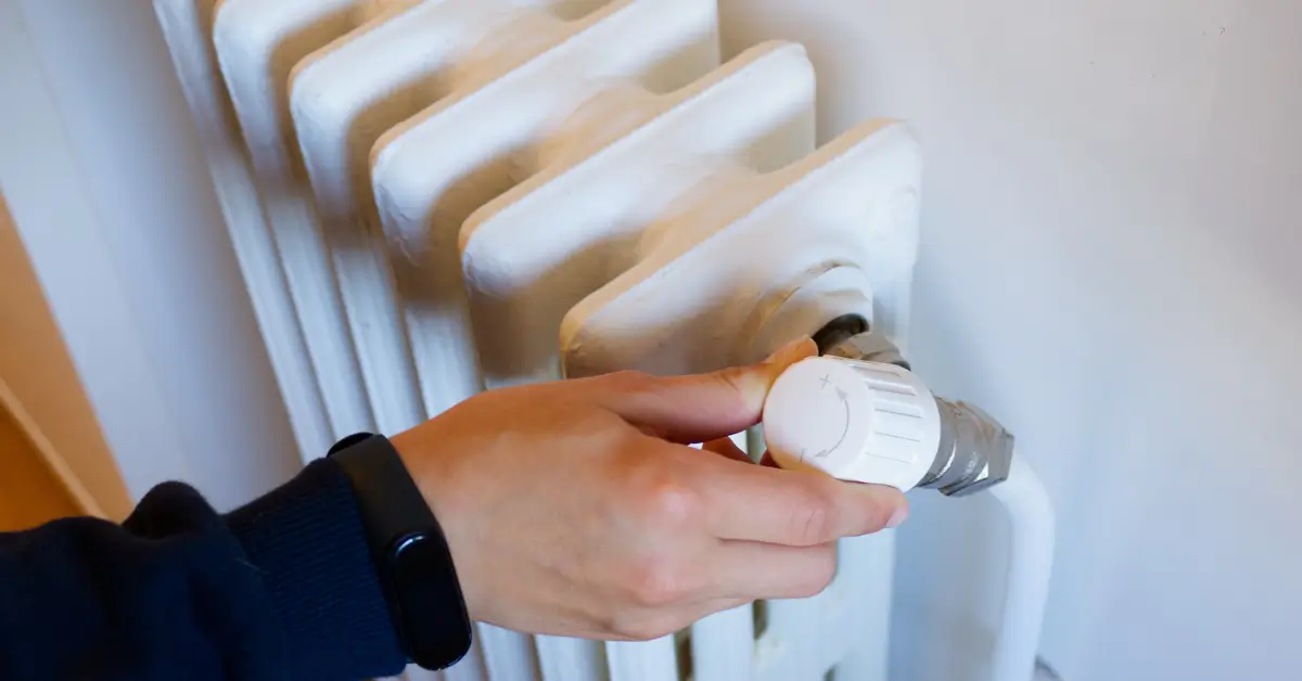 A person’s hand adjusting the temperature control knob on a white radiator heater mounted against a wall.
